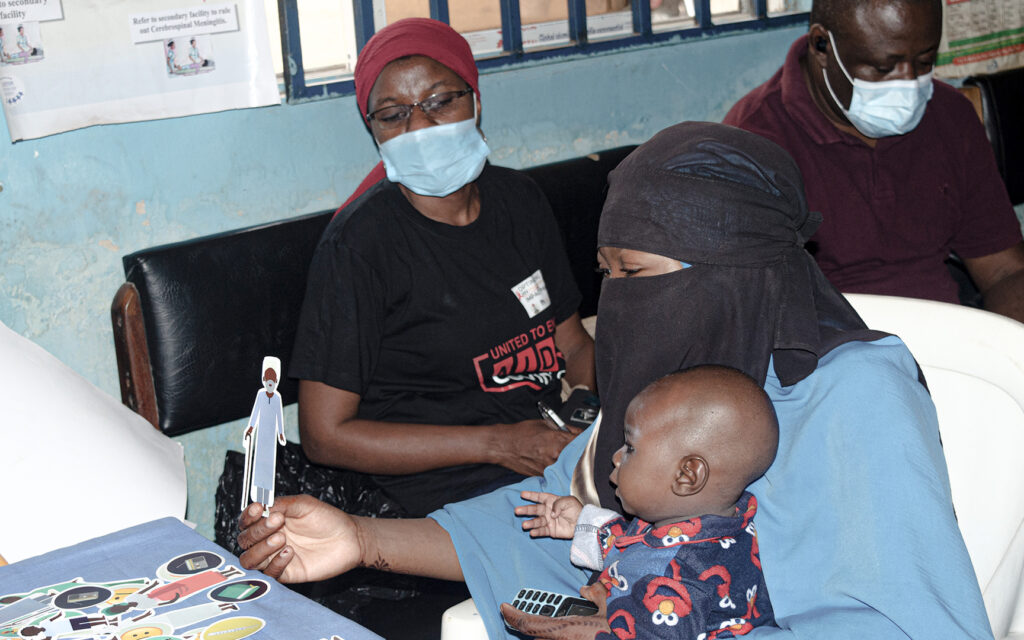 Woman wearing a burqa holds a paper doll depicting a community elder. She has a baby in her lap and other prompts in front of her. A Niger team member is sitting on a bench at the side, listening closely to the woman's explanation.